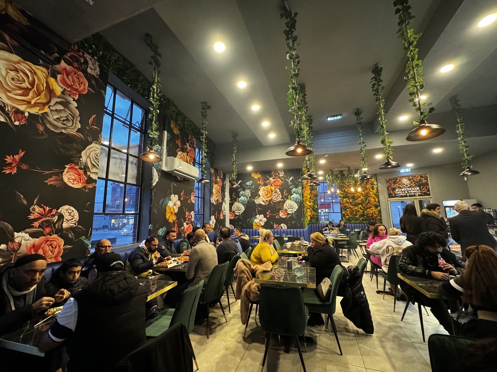Inside Kohinoor Grill and Thrill: floral wallpaper, green velvet chairs, trailing ivy and warm pendant lighting over a busy dining room